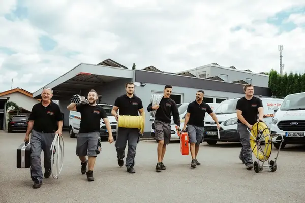 Eine Gruppe Elektriker läuft mit Werkzeug und Kabeln vor Firmenfahrzeugen auf dem Betriebshof.