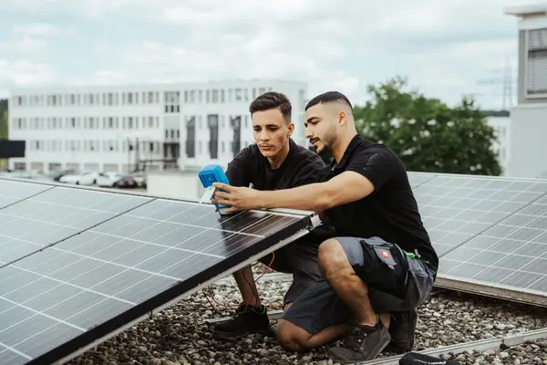 Zwei Techniker prüfen mit einem Messgerät Solaranlagen auf einem Flachdach bei Tageslicht.